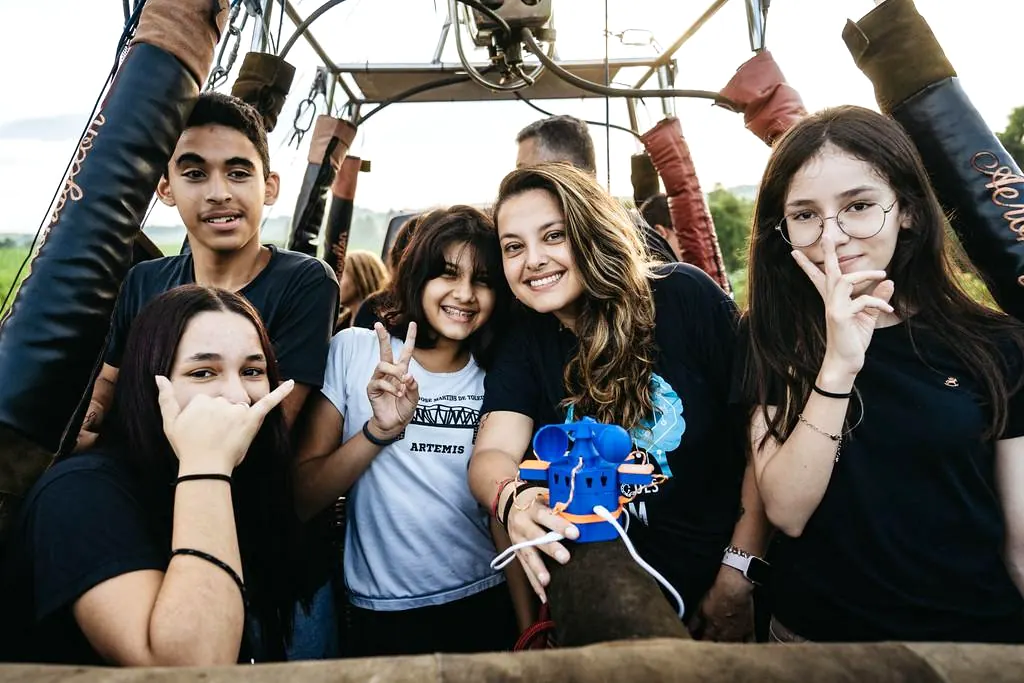 Alunos da E.E. Prof. José Martins de Toledo, de Artemis, sorriem no cesto de um balão de ar quente segurando a estação meteorológica azul impressa em 3D.