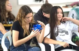 Estudantes sentadas observam a estação meteorológica azul, impressa em 3D, que uma delas segura nas mãos.